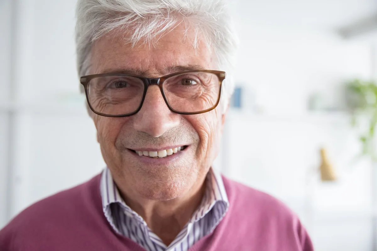 An older man in glasses and a soft pink sweater, smiling broadly at the camera