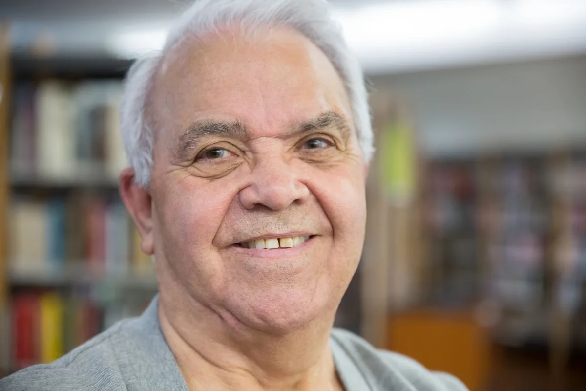 An older man with white hair smiling warmly, sitting in a sunlit room lined with books